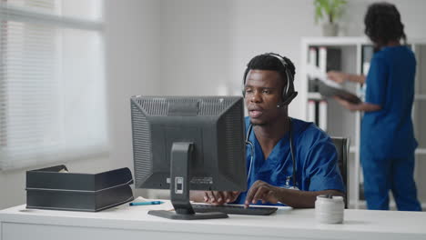 Afroamerikanischer-Männlicher-Arzt-Oder-Krankenschwester-Mit-Headset-Und-Computer,-Der-Im-Krankenhaus-Arbeitet.-Junger-Professioneller-Therapeut,-Arzt,-Der-Den-Kunden-Berät,-Indem-Er-Fernkommunikation-Nutzt-Und-über-Die-Webcam-Spricht